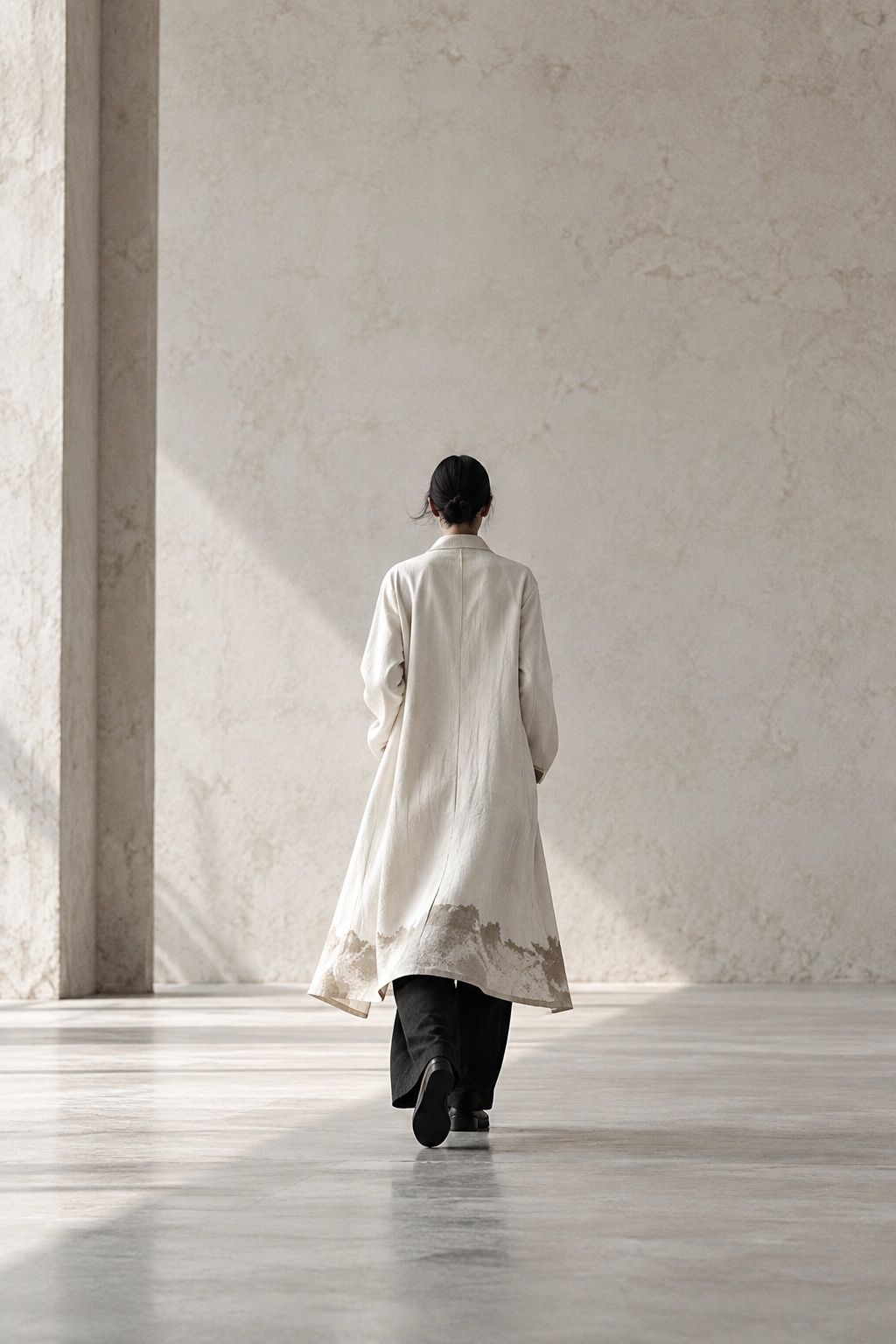 Model walking away from camera in a long bone linen coat with a pale tide mark at the hem, black trousers, bare concrete floor