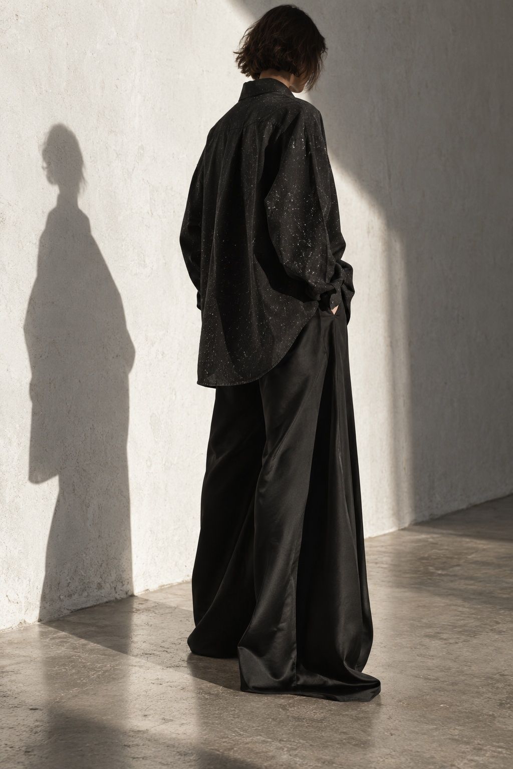 Model in an oversized black shirt and wide black trousers, face turned away, standing in soft side light